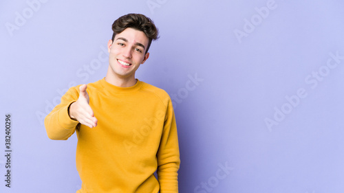 Young caucasian man isolated on purple background stretching hand at camera in greeting gesture. © Asier