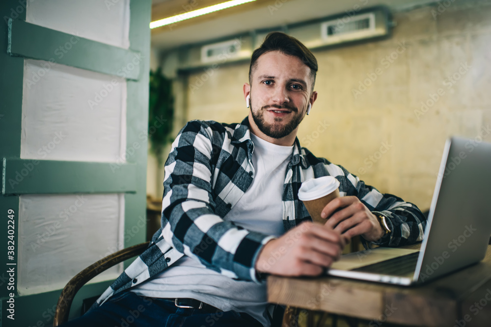 Smiling male hipster with takeaway coffee and laptop in cafe