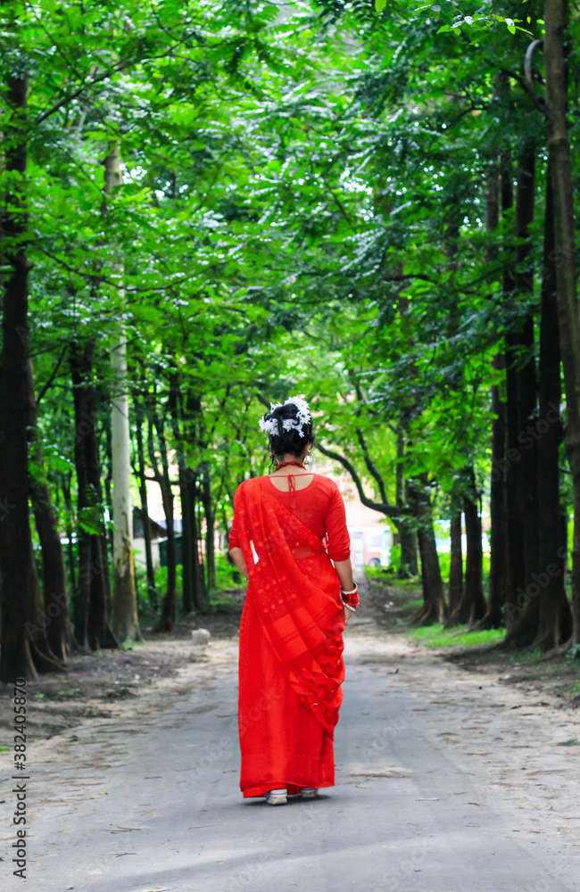 Young woman wearing red saree walking on green asphalt road. The girl walks along the path in the woods to the light. Chattogram/ 2020.