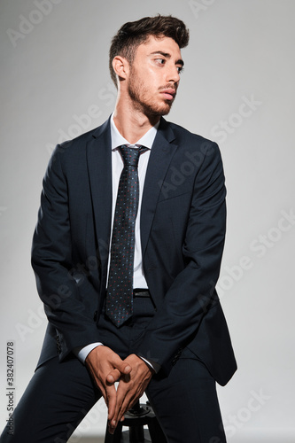 Young man portrait seated in a stool wearing a dark blue suit looking to his left.