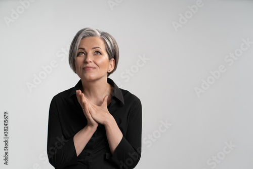 Middle-aged woman has folded her palms and looks up dreamily. Woman with gray hair in a black shirt on an isolated background. High quality photo.