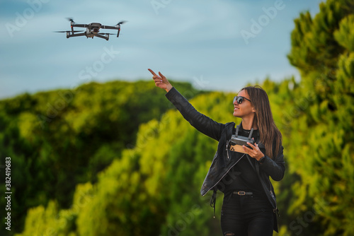 Cute modern smiling woman pilot in dark stylish clothes flying drone in park at sunset. Outdoors hobby.