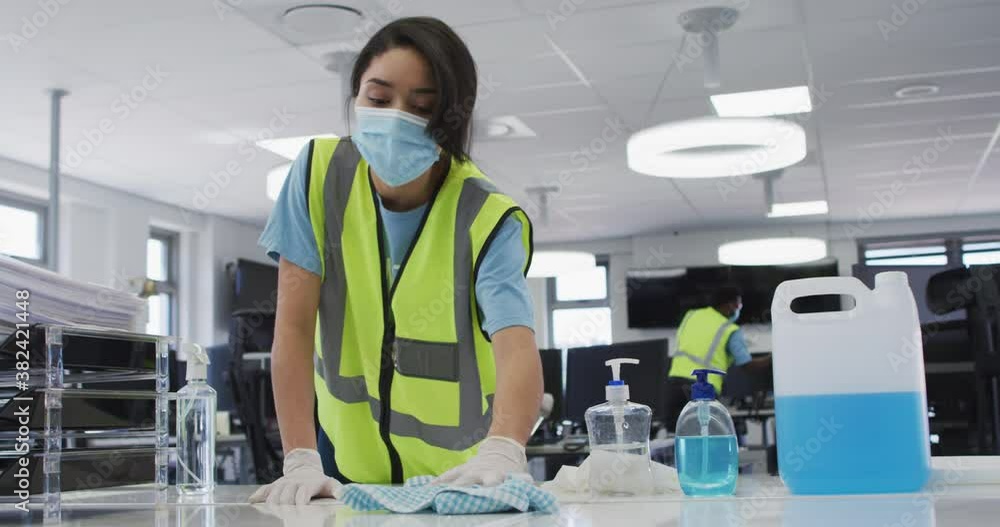 Mixed race woman wearing hi vis vest and face mask cleaning the office using disinfectant Mixed race woman wearing hi vis vest and face mask cleaning the office using disinfectant