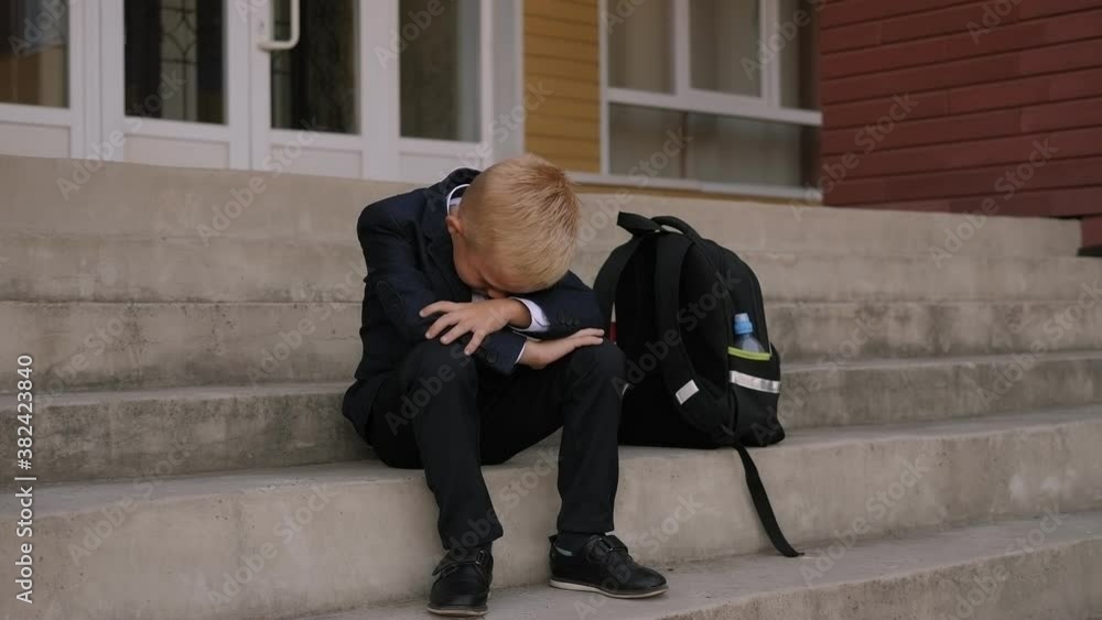 A distressed boy covers his face with his hands and cries while sitting on the school stairs, his backpack is next to him. A distressed boy covers his face with his hands and cries while sitting on the school stairs, his backpack is next to him.