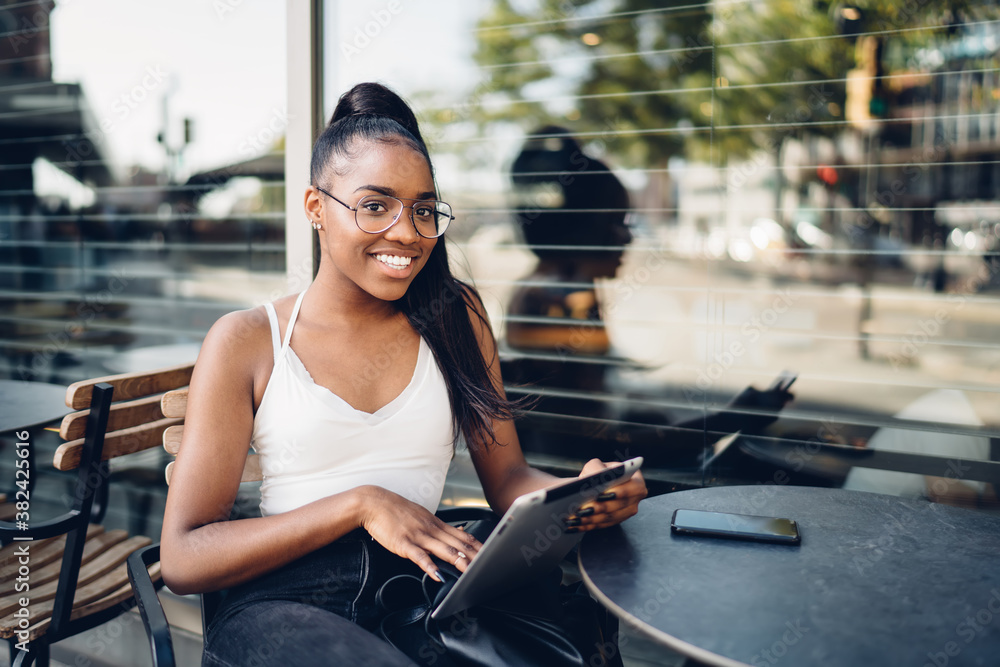 Fototapeta premium Portrait of cheerful dark skinned hipster girl in eyewear using digital tablet for searching information, happy african american woman blogger resting on free time on cafe terrace with touchpad