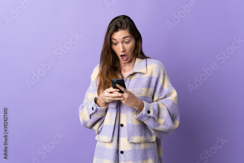 Young Ireland woman isolated on purple background looking at the camera while using the mobile with surprised expression