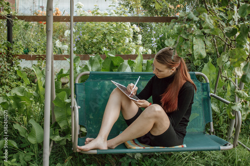 Foto Cute teenage girl artist with red hair sits in the garden on a swing and draws sketches on a tablet