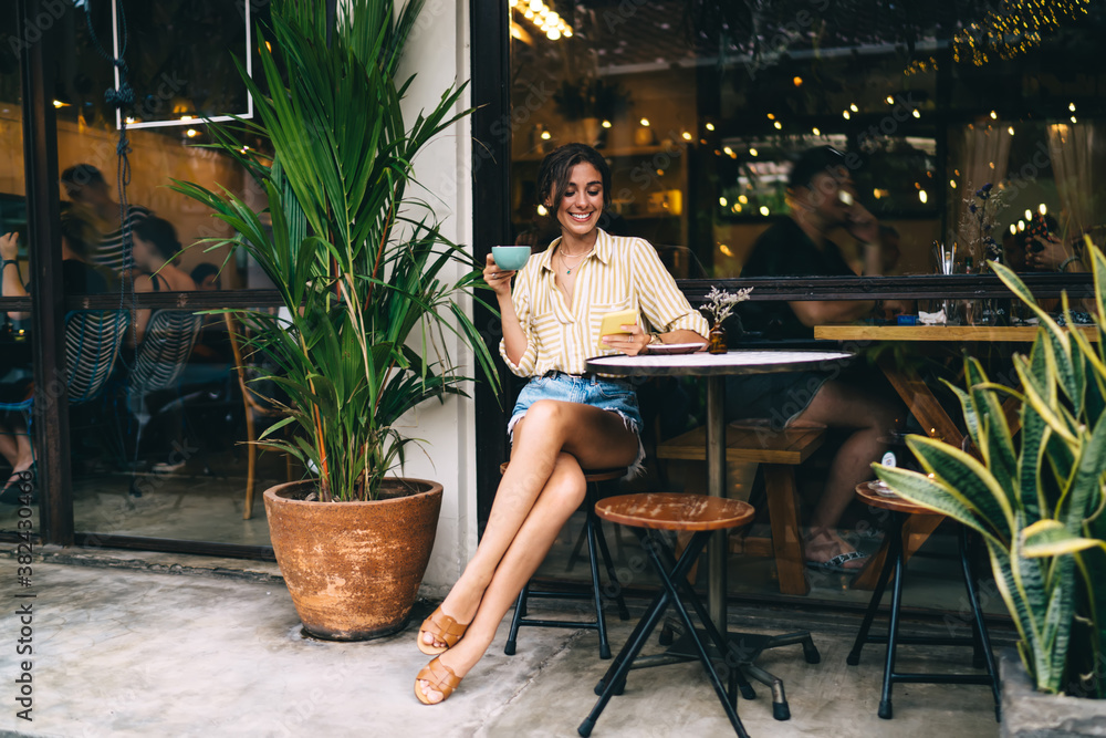 Cheerful young ethnic woman sitting in sidewalk cafe with smartphone Cheerful young ethnic woman sitting in sidewalk cafe with smartphone