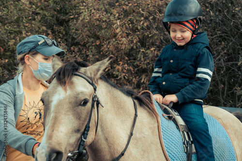 Happy kid boy riding a horse training with instructor. The boy learns to ride a horse. Hippotherapy for the development of the baby. A child in a warm suit and helmet for safety. quarantine mask