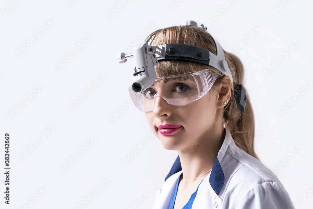 Confident ENT doctor wearing surgical headlight head light and protective glasses. Closeup portrait of female otolaryngologist or head and neck surgeon on light grey background.