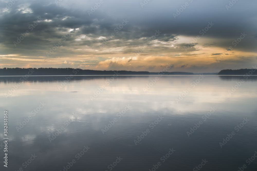 The smooth surface of the water reflects the dramatic sky. A dark strip of forest can be seen in the background.