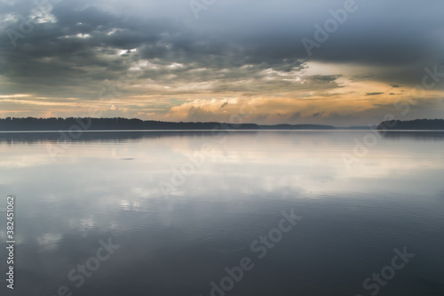 The smooth surface of the water reflects the dramatic sky. A dark strip of forest can be seen in the background.