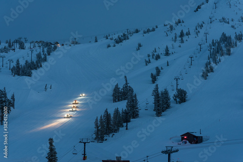 Foto Snow machines with headlights on grooming ski slopes at twilight/dusk on a mountain with trees and a cabin