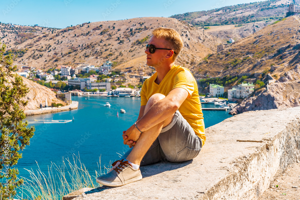 Balaklava / Crimea - 20 Sep 2020: A young man in bright clothes looks at the Yachts standing in the Bay with a view of the city and the azure water