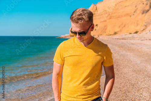 A young man in bright clothes and sunglasses walks on a sandy beach near the sea