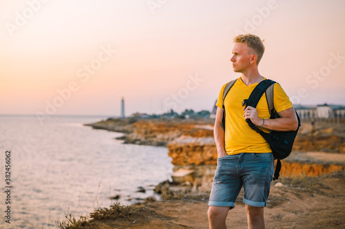 Fototapeta Naklejka Na Ścianę i Meble -  A young man with a backpack looks at the sunset with a lighthouse in the background