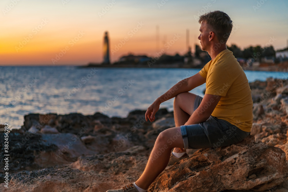 A young man sits thoughtfully on a rock and looks at the sunset, with a lighthouse by the sea in the background A young man sits thoughtfully on a rock and looks at the sunset, with a lighthouse by the sea in the background