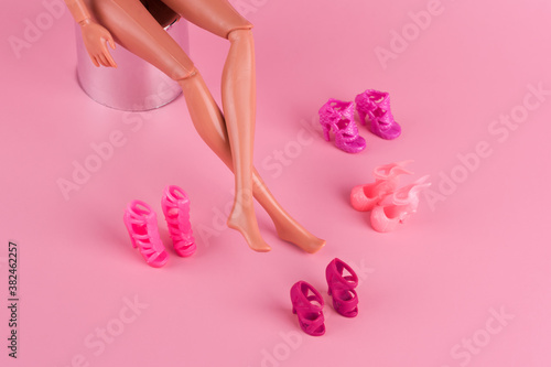 Doll with long skinny legs and stylish shoes on pink background. Plastic doll trying on high heels shoes. Woman choosing new shoes, deciding which pair to buy. Fashion and shopping concept.