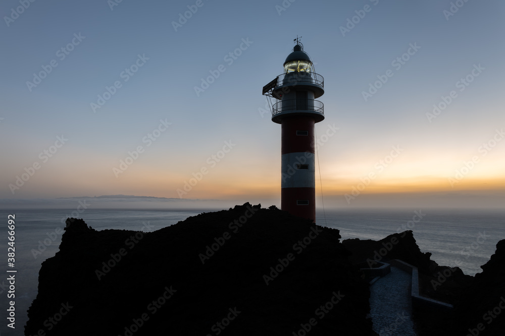 The lighthouse Punta de Teno in the far west of the Atlantic island of Tenerife at sunset. The neighboring island of La Gomera can be seen in the background behind clouds.