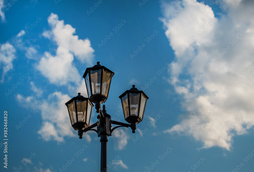 Old vintage black decorative lantern with clear glass on pillar. Three street lamps on one pole. Sunny blue sky with white clouds