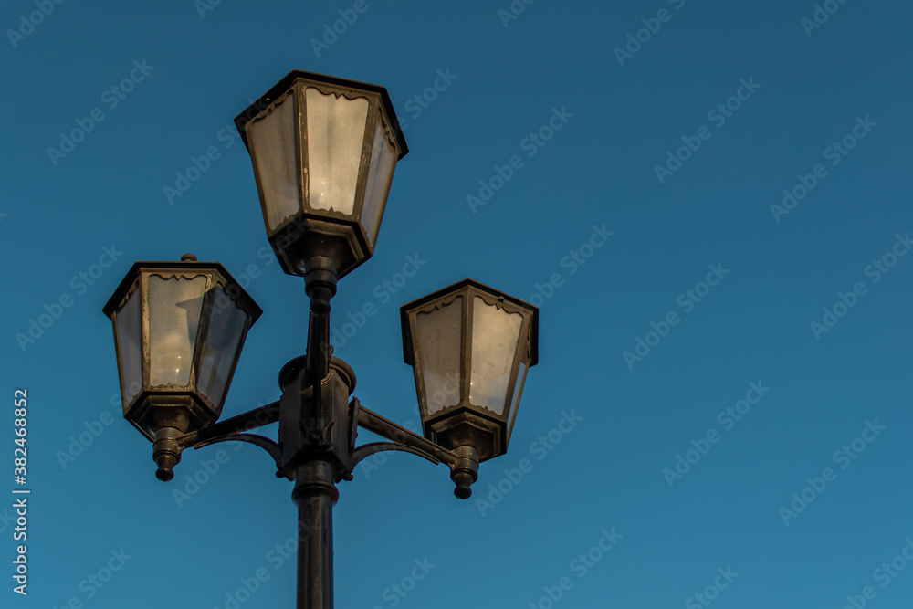 Old vintage black decorative lantern with clear glass on pillar. Three street lamps on one pole in sunset light. Blue sky