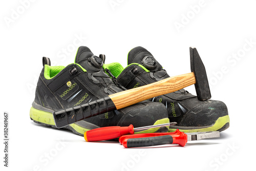 Pair of black safety shoes with with green lining along with a hammer with wooden handle, a red pliers and a red Phillips screwdriver. With a white background.