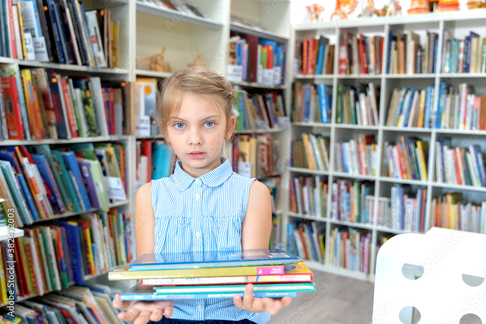 Little girl holds stack of books with fairy tales in children's library. Special reading kids room. Shelves with bright interesting, educational books about animals.Table,chair Little girl holds stack of books with fairy tales in children's library. Special reading kids room. Shelves with bright interesting, educational books about animals.Table,chair