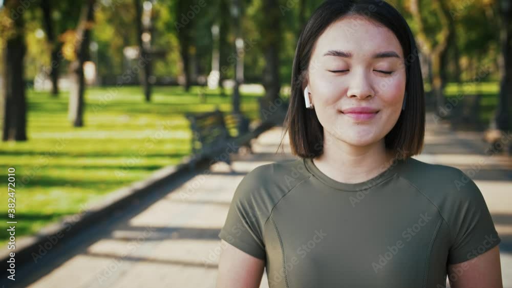 Adorable asian woman in wireless headphones smiling to camera in city park, tracking shot Adorable asian woman in wireless headphones smiling to camera in city park, tracking shot