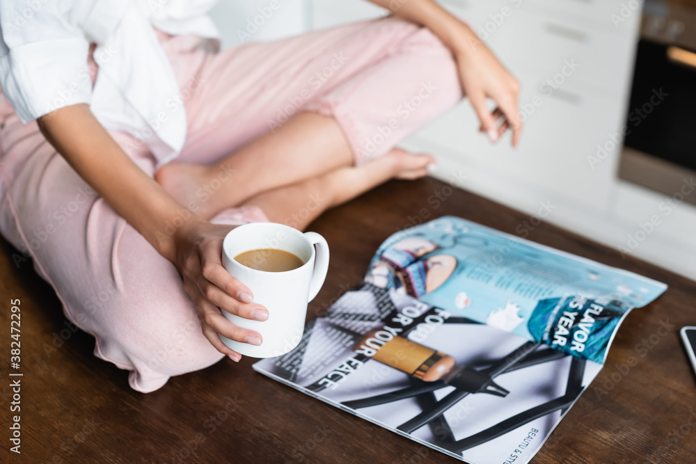 cropped view of woman holding cup of coffee near magazine on table cropped view of woman holding cup of coffee near magazine on table