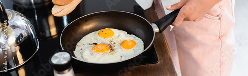 cropped view of woman holding kitchen tongs near eggs on frying pan, panoramic shot