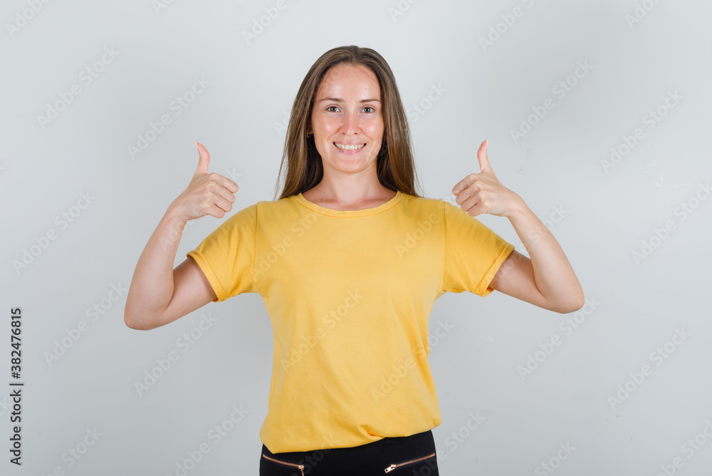 Young woman showing thumbs up in yellow t-shirt, pants and looking cheery. front view. Young woman showing thumbs up in yellow t-shirt, pants and looking cheery. front view.