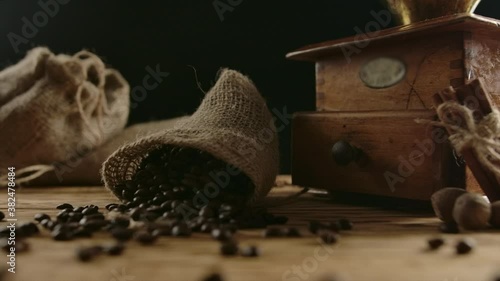 Suddenly falling jute bag with roasted coffee beans. Grains scattering on the wooden table in slow motion. Vintage coffee grinder, cinnamon and nutmeg on background