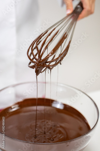 Closeup stirring milk melted chocolate in glass bowl with whisk isolated on white background. Chocolatier make premium hand-crafted chocolate. Candy making, pastry production, bakery, dessert concept