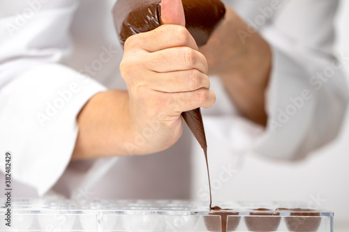 Close-up chocolatier pours dark chocolate into sweets mold. Chef in white apron using pastry bag filling out plastic mold with hot melted chocolate. Concept for making homemade chocolate candy dessert