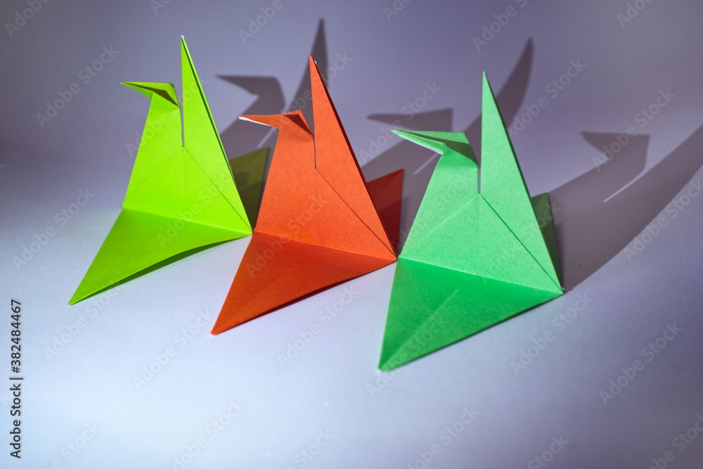 three origami in the shape of a tsuru, folding on paper with green and red colors, casting their shadows on a light background. three origami in the shape of a tsuru, folding on paper with green and red colors, casting their shadows on a light background.