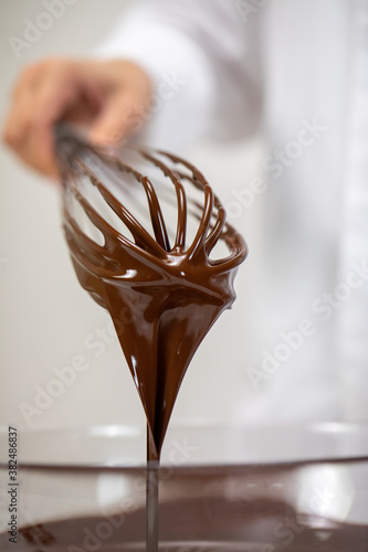 Closeup pouring tasty dark melted couverture chocolate on whisk isolated on white background. Chocolatier making premium hand-crafted chocolate. Candy making, pastry bakery production, dessert concept
