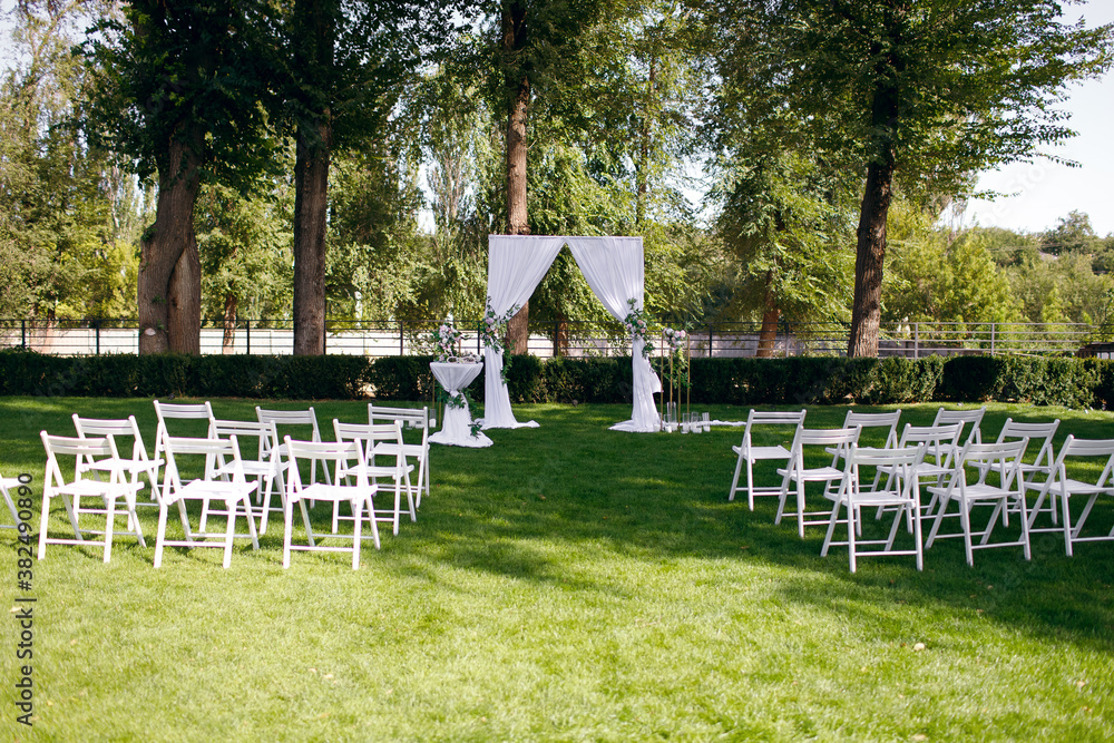 a wedding arch decorated with natural flowers stands on a green lawn in the forest, white chairs for guests a wedding arch decorated with natural flowers stands on a green lawn in the forest, white chairs for guests