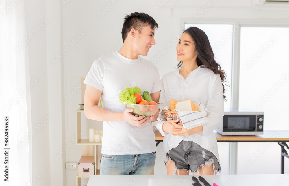 asian man and asian woman cooking organic sandwich in kitchen room, they feeling happy and smile in breakfast time, they holding vegetables and breads