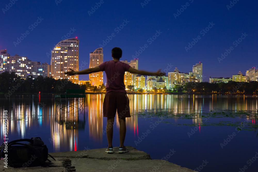 A guy stands on the banks of the Dnieper river against the background of a bright night embankment