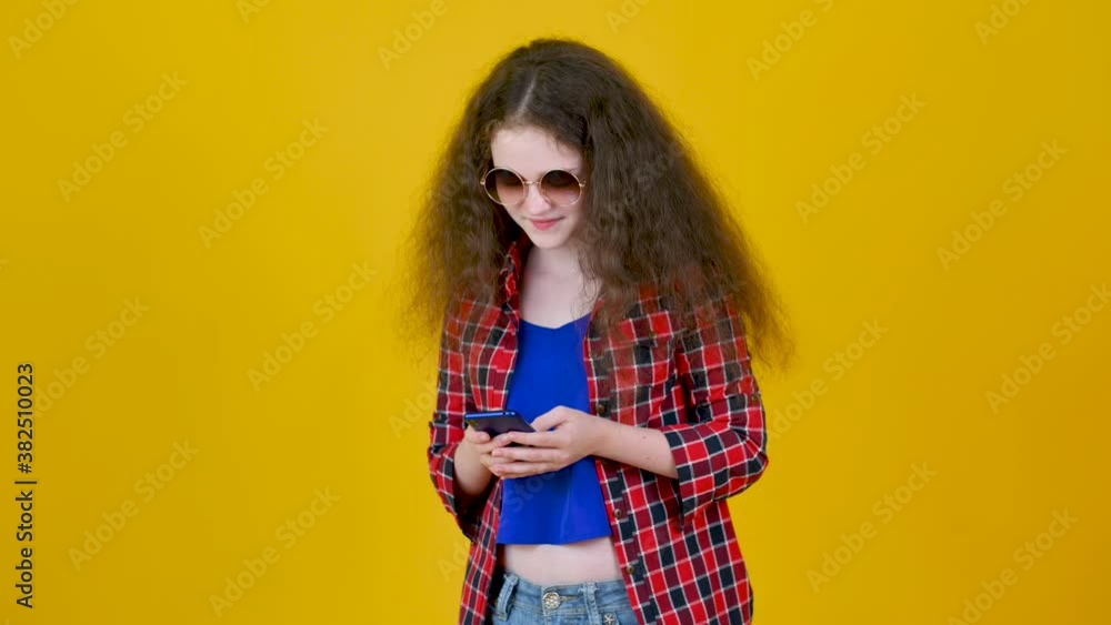 Young girl in glasses with curly hair using smartphone texting sharing messages on social media on yellow studio background. Young girl chatting on phone Young girl in glasses with curly hair using smartphone texting sharing messages on social media on yellow studio background. Young girl chatting on phone