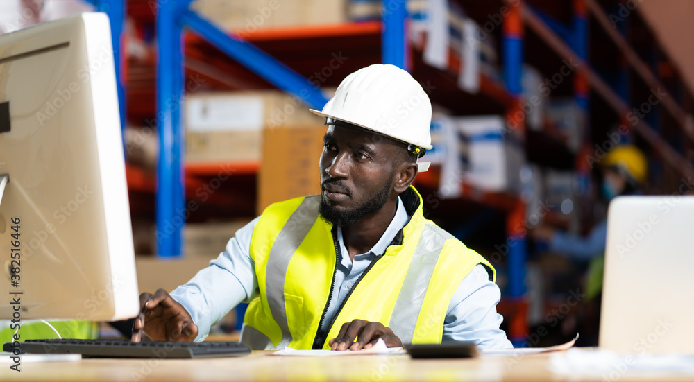 middle aged African American warehouse worker working on personal computer and preparing a shipment in large warehouse distribution centre middle aged African American warehouse worker working on personal computer and preparing a shipment in large warehouse distribution centre