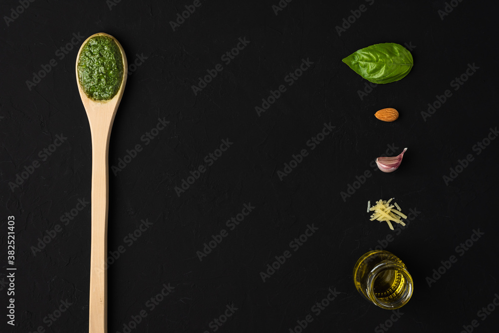 Basil pesto sauce on a wooden spoon with a basil leaf, almond, garlic, parmesan cheese and olive oil as ingredients on a black background with space for text. Basil pesto sauce on a wooden spoon with a basil leaf, almond, garlic, parmesan cheese and olive oil as ingredients on a black background with space for text.