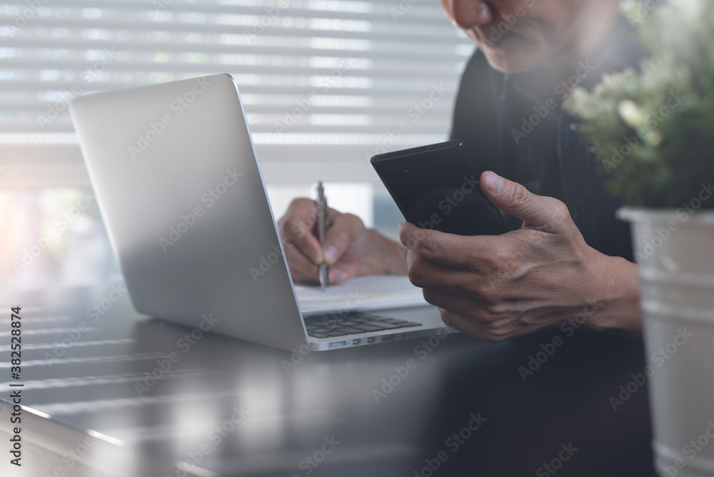 Man using mobile phone and writing on notepad while working on laptop computer in office Man using mobile phone and writing on notepad while working on laptop computer in office