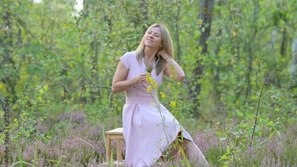 A cute blonde girl in a pink dress is resting on a green meadow. The girl is holding heather flowers in her hands and smiling. A cute blonde girl in a pink dress is resting on a green meadow. The girl is holding heather flowers in her hands and smiling.