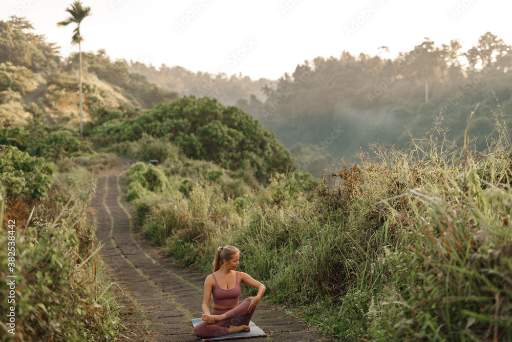 Young woman doing stretching or yoga exercise on a mat, nature background. Yoga training in the park. Sport and fitness concept outdoor