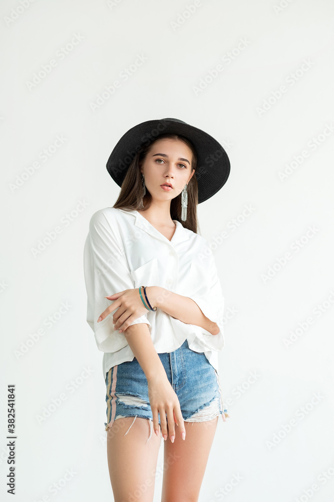 Summer trend. Casual style. Pretty young woman in white shirt jeans shorts and straw black hat combining vintage earrings looking at camera isolated on neutral. Fashion model. Romantic look Summer trend. Casual style. Pretty young woman in white shirt jeans shorts and straw black hat combining vintage earrings looking at camera isolated on neutral. Fashion model. Romantic look