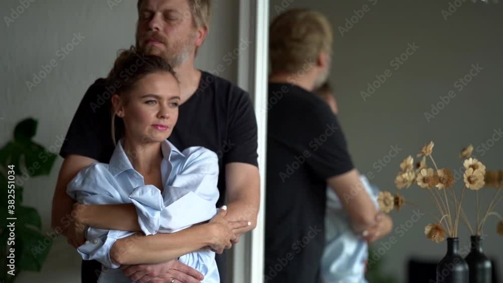 Reflection in the mirror of a couple in love, gently embracing in a modern home room. A gorgeous woman and a handsome man hugging her from behind. Reflection in the mirror of a couple in love, gently embracing in a modern home room. A gorgeous woman and a handsome man hugging her from behind.