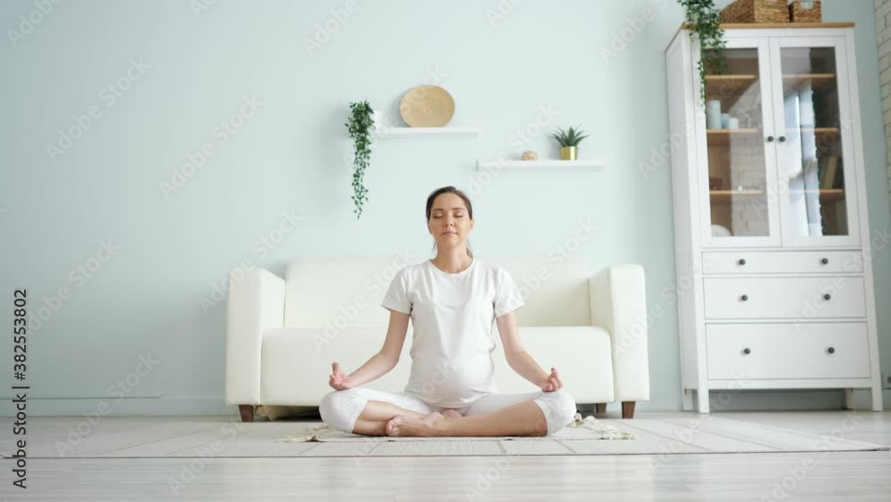 Smiling young pregnant lady in white meditates sitting in padmasana yoga position on floor against designer furniture in spacious room at home, camera zoom in Smiling young pregnant lady in white meditates sitting in padmasana yoga position on floor against designer furniture in spacious room at home, camera zoom in
