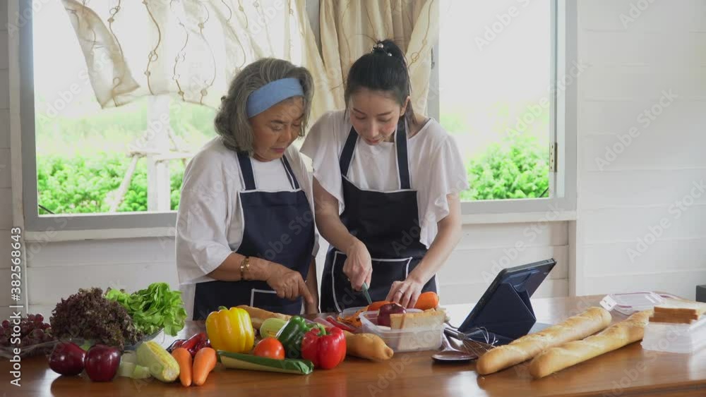 happy asian young daughter and senior mother cooking online class on tablet together making fresh vegetables food in kitchen at home happy asian young daughter and senior mother cooking online class on tablet together making fresh vegetables food in kitchen at home