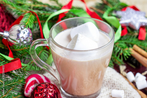 Hot chocolate Christmas and New Year celebration cocoa drink in a glass cup with fir tree branches, holiday decorations and sweet red striped lollipops on wooden background.